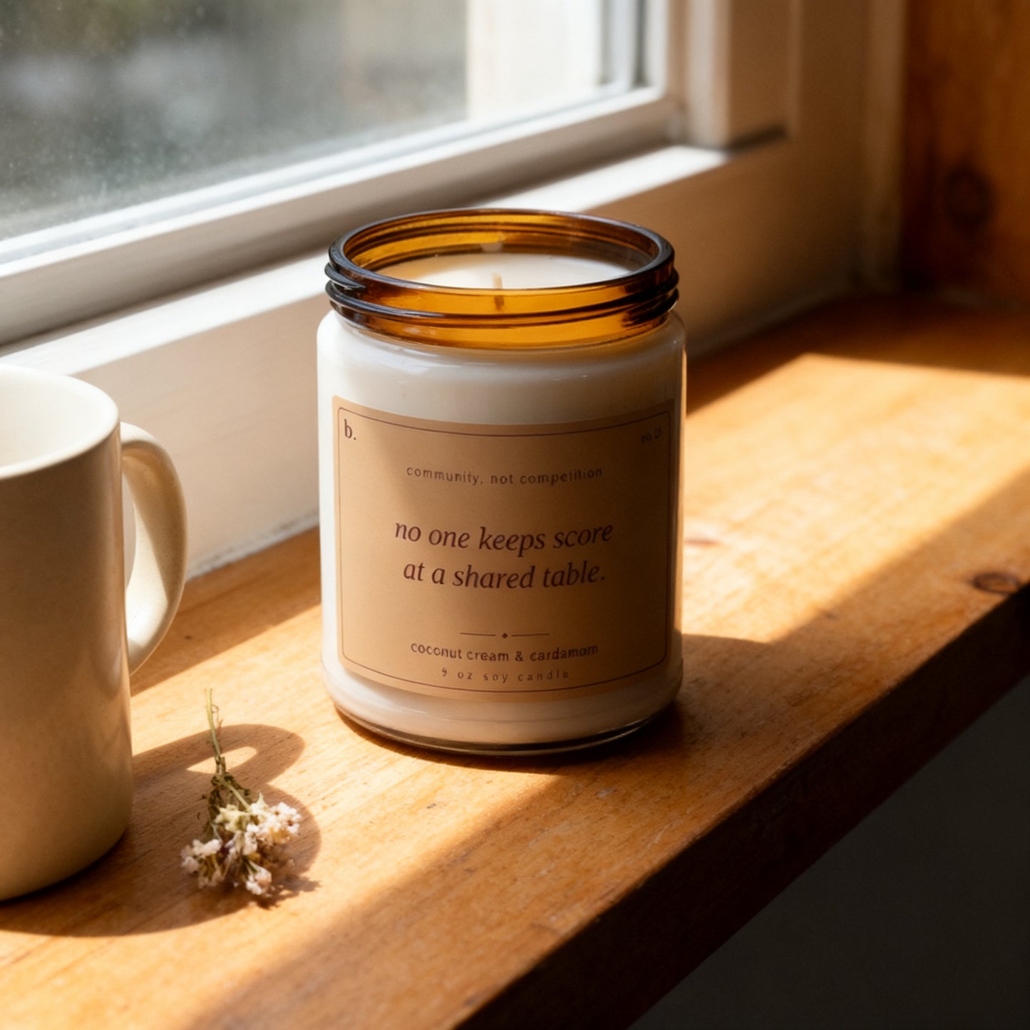 b.together coconut cream and cardamom candle on a sunlit windowsill with a ceramic mug and dried flowers